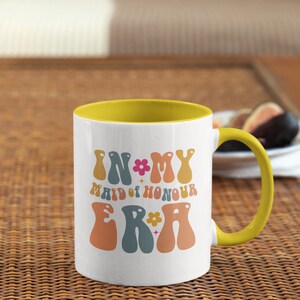 May include: White ceramic mug with a yellow rim and handle. The mug has a retro design with the text "In My Maid of Honour Era" in colorful, wavy letters.