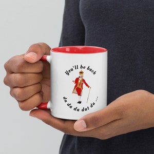 May include: White ceramic mug with a red rim. The mug features a cartoon illustration of a king in a red and gold costume, singing "You'll be back da da da dat da..."