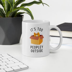 May include: White ceramic mug with a black and orange illustration of a cat in a box. The text "It's too peopley outside" is printed on the mug.