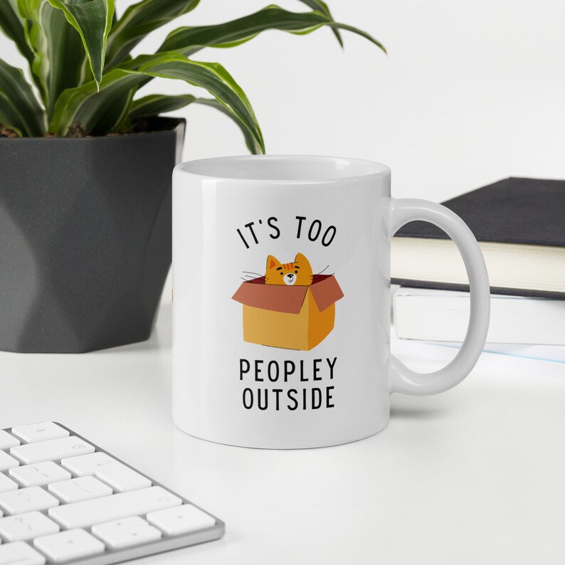 May include: White ceramic mug with a black and orange illustration of a cat in a box. The text "It's too peopley outside" is printed on the mug.