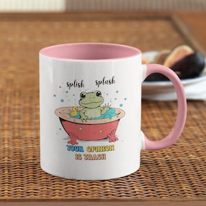 May include: White ceramic mug with a pink interior and handle. The mug features a cartoon frog in a bathtub with the text "splish splash" and "YOUR OPINION IS TRASH". A yellow rubber duck is also in the bathtub.