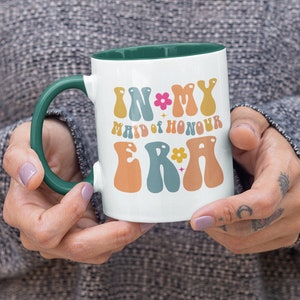 May include: White ceramic mug with a green rim and handle. The mug has a colorful graphic that says "In My Maid of Honour Era" with flowers and stars.