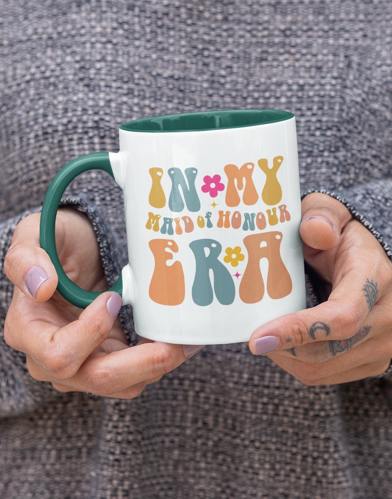 May include: White ceramic mug with a green rim and handle. The mug has a colorful graphic that says "In My Maid of Honour Era" with flowers and stars.