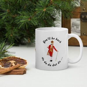 May include: White ceramic mug with a black and white illustration of a man in a red and gold costume with a crown. The text "You'll be back da da da dat da..." is printed on the mug.