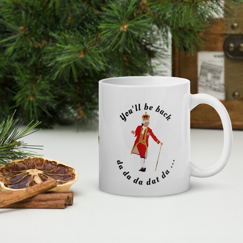May include: White ceramic mug with a black and white illustration of a man in a red and gold costume with a crown. The text "You'll be back da da da dat da..." is printed on the mug.