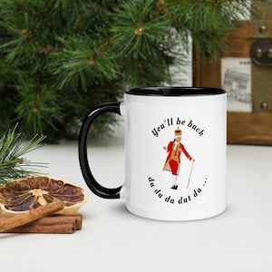 May include: White ceramic mug with a black rim and handle. The mug features a cartoon illustration of a king in a red and gold uniform, holding a cane and singing. The text "You'll be back da da da da dat da..." is printed on the mug.