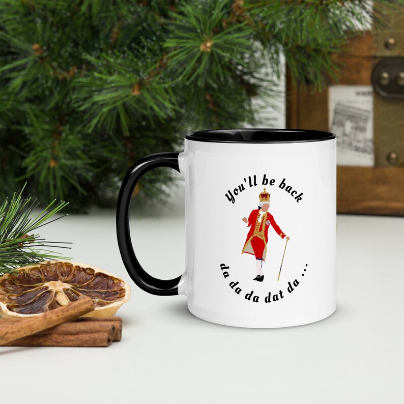 May include: White ceramic mug with a black rim and handle. The mug features a cartoon illustration of a king in a red and gold uniform, holding a cane and singing. The text "You'll be back da da da da dat da..." is printed on the mug.