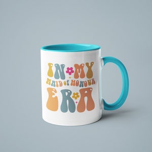 May include: White ceramic mug with a turquoise rim and handle. The mug has a colorful graphic that says "In My Maid of Honour Era" with flowers and stars.