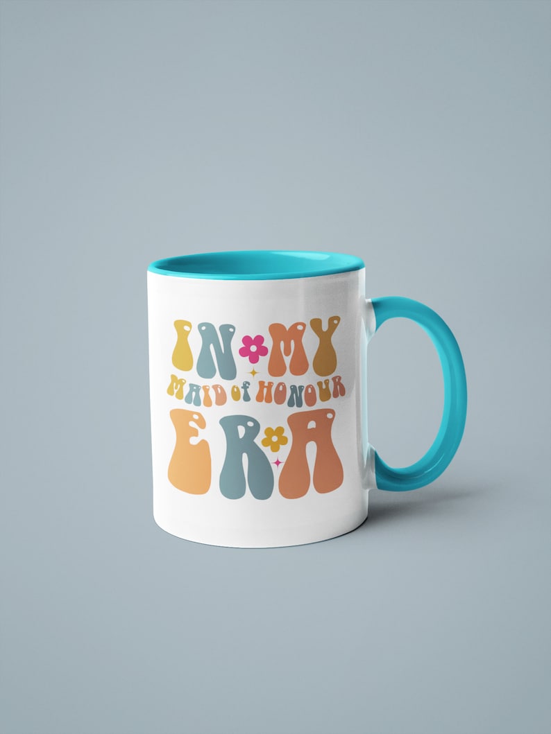 May include: White ceramic mug with a turquoise rim and handle. The mug has a colorful graphic that says "In My Maid of Honour Era" with flowers and stars.