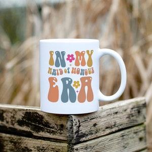 May include: White ceramic mug with the text "In My Maid of Honour Era" in colorful, retro-style lettering with flowers and stars.