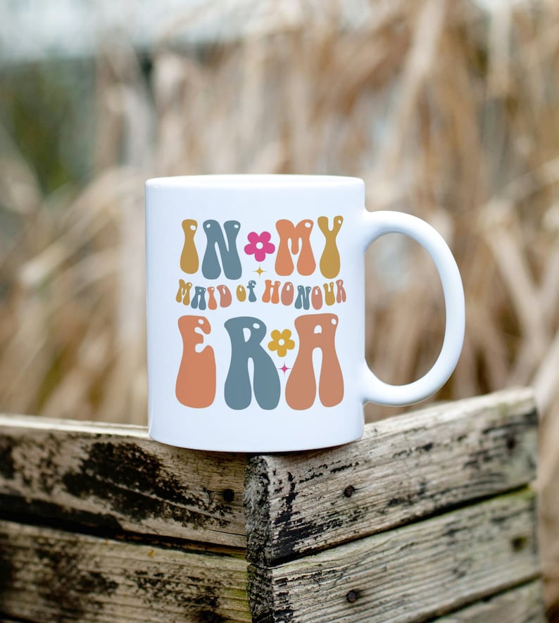 May include: White ceramic mug with the text "In My Maid of Honour Era" in colorful, retro-style lettering with flowers and stars.
