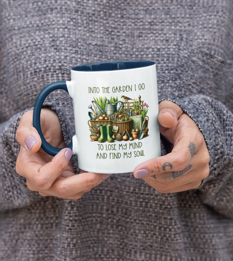 May include: White ceramic mug with a blue rim and handle. The mug features a colorful illustration of gardening tools and the text "Into the garden I go to lose my mind and find my soul."