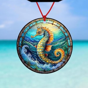 Peut inclure: Une boule de Noël ronde en verre avec un motif de vitrail représentant un hippocampe bleu, jaune et orange nageant dans une mer bleue avec des vagues blanches. La boule est suspendue à une ficelle rouge.