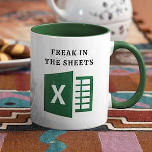Può includere: Una tazza da caffè bianca con un bordo e un manico verdi. La tazza ha un logo Microsoft Excel verde e il testo "Freak in the sheets".