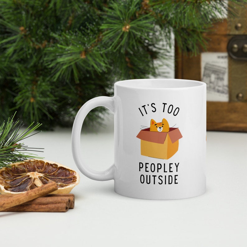 May include: White ceramic mug with a black and orange illustration of a cat in a box. The text "It's too peopley outside" is printed on the mug.