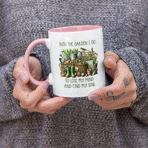 May include: White ceramic mug with a pink rim and handle. The mug features a colorful illustration of gardening tools, plants, and a bird. The text "Into the garden I go to lose my mind and find my soul" is printed on the mug.