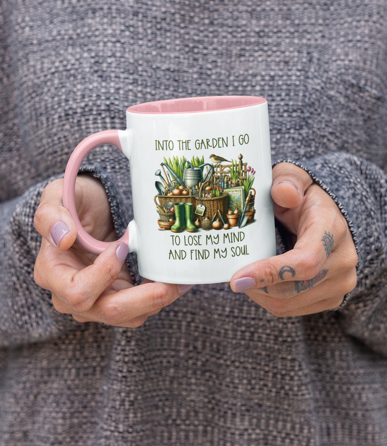 May include: White ceramic mug with a pink rim and handle. The mug features a colorful illustration of gardening tools, plants, and a bird. The text "Into the garden I go to lose my mind and find my soul" is printed on the mug.