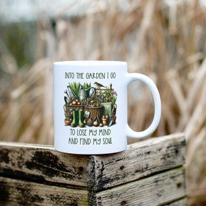 May include: White ceramic mug with a colorful illustration of gardening tools, plants, and a bird. The text "Into the garden I go to lose my mind and find my soul" is printed on the mug.