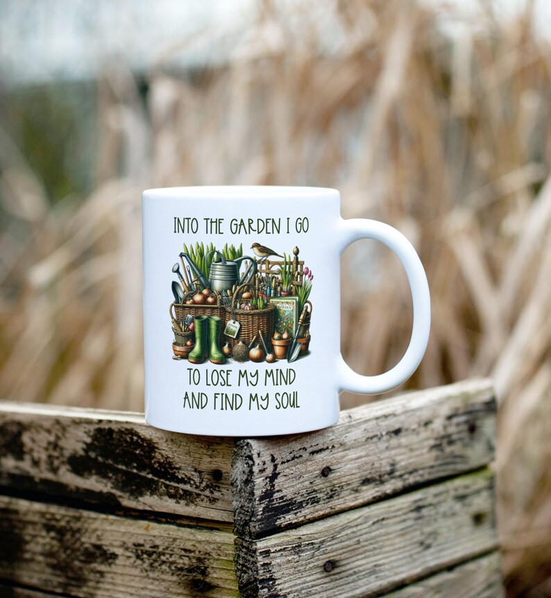 May include: White ceramic mug with a colorful illustration of gardening tools, plants, and a bird. The text "Into the garden I go to lose my mind and find my soul" is printed on the mug.