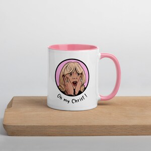 May include: White ceramic mug with a pink interior and handle. The mug features a cartoon illustration of a surprised woman with the text "Oh my Christ!" in black. The mug is on a wooden surface.