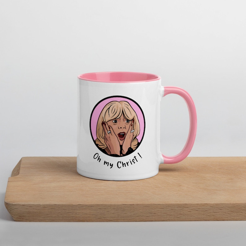 May include: White ceramic mug with a pink interior and handle. The mug features a cartoon illustration of a surprised woman with the text "Oh my Christ!" in black. The mug is on a wooden surface.