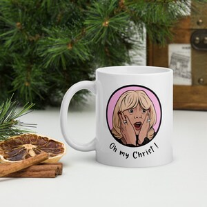 May include: White ceramic mug featuring a cartoon illustration of a surprised blonde woman with the text "Oh my Christ!". The design is set within a pink circle. The mug has a curved handle.