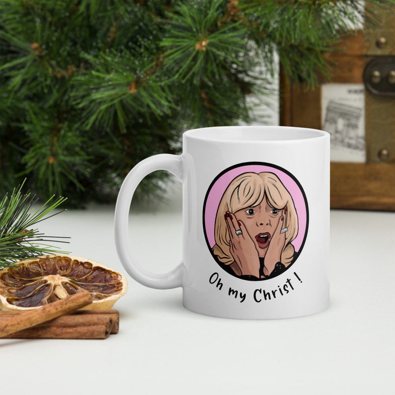 May include: White ceramic mug featuring a cartoon illustration of a surprised blonde woman with the text "Oh my Christ!". The design is set within a pink circle. The mug has a curved handle.