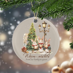 Puede incluir: Adorno navideño redondo con una escena invernal que presenta tres conejos cantando villancicos cerca de un árbol de Navidad y una farola. El adorno tiene el texto "Warm wishes from our family to yours".