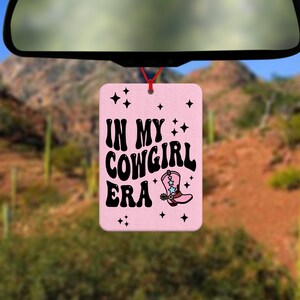 Pode incluir: Um ambientador retangular rosa com o texto "IN MY COWGIRL ERA" em preto e um desenho de uma bota de cowboy. Est&aacute; pendurado por um fio vermelho, com uma paisagem des&eacute;rtica ao fundo.
