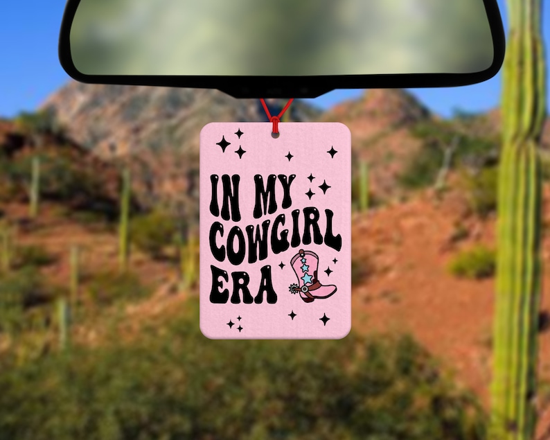 Pode incluir: Um ambientador retangular rosa com o texto "IN MY COWGIRL ERA" em preto e um desenho de uma bota de cowboy. Est&aacute; pendurado por um fio vermelho, com uma paisagem des&eacute;rtica ao fundo.