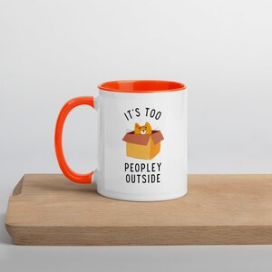 May include: White ceramic mug with an orange rim and handle. The mug has a cartoon cat in a box with the text "It's too peopley outside".