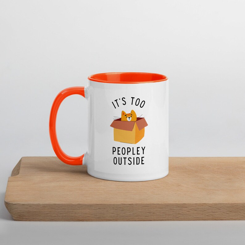 May include: White ceramic mug with an orange rim and handle. The mug has a cartoon cat in a box with the text "It's too peopley outside".