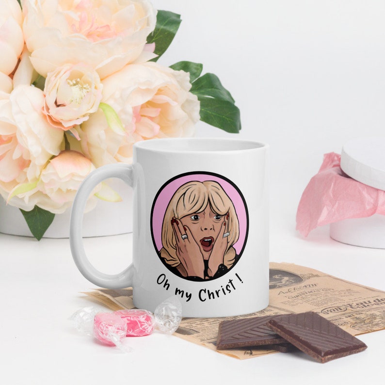 May include: White ceramic mug featuring a cartoon illustration of a surprised woman with blonde hair and the text "Oh my Christ!". The design is set within a pink circle. The mug has a curved handle and is surrounded by flowers and chocolate.