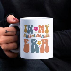 May include: White ceramic mug with the text "In My Maid of Honour Era" in colorful retro font with a flower and star design.
