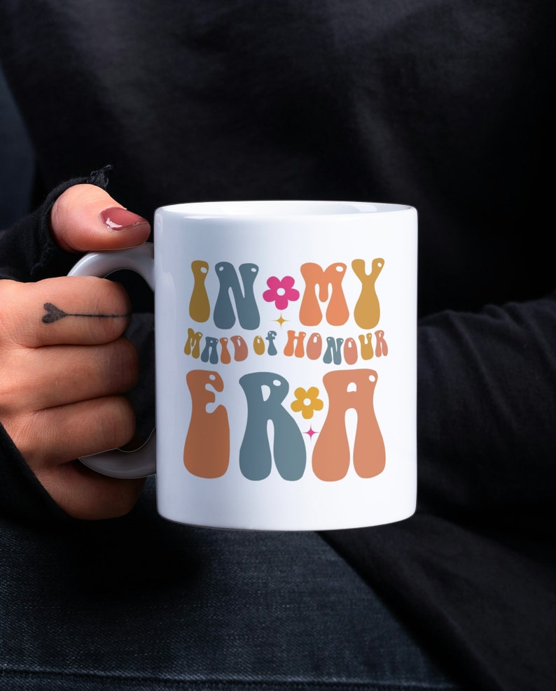 May include: White ceramic mug with the text "In My Maid of Honour Era" in colorful retro font with a flower and star design.