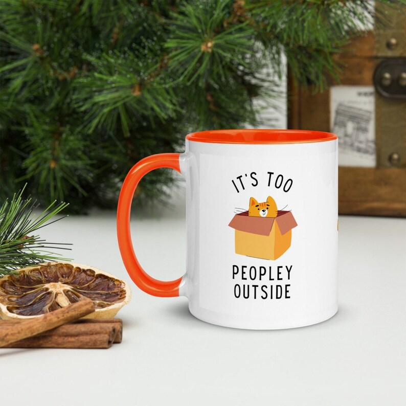May include: White ceramic mug with an orange rim and handle. The mug has a graphic of an orange cat in a box with the text "It's too peopley outside".
