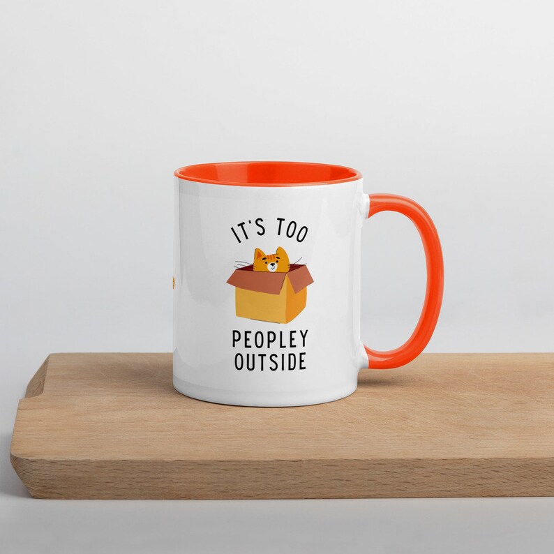 May include: White ceramic mug with an orange rim and handle. The mug features a cartoon cat in a box with the text "It's too peopley outside".