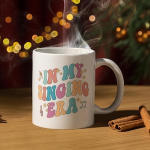 Könnte beinhalten: Eine weiße Keramiktasse mit einem bunten Retro-Design, auf dem "In My Singing Era" mit Musiknoten steht. Die Tasse ist mit einem dampfenden Getränk gefüllt und steht auf einer Holzoberfläche.