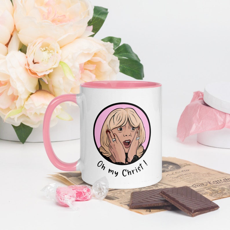 May include: White ceramic mug with a pink interior and handle. The mug features a cartoon illustration of a surprised woman with the text "Oh my Christ!" in black. The mug is surrounded by flowers, candy, and chocolate.