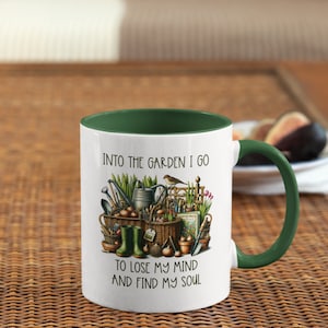 May include: White ceramic mug with a green rim and handle. The mug features a colorful illustration of gardening tools, plants, and a bird. The text "Into the garden I go to lose my mind and find my soul" is printed on the mug.