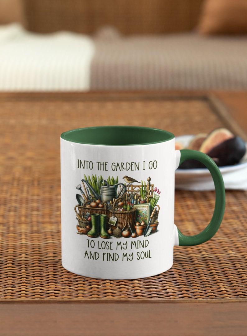 May include: White ceramic mug with a green rim and handle. The mug features a colorful illustration of gardening tools, plants, and a bird. The text "Into the garden I go to lose my mind and find my soul" is printed on the mug.