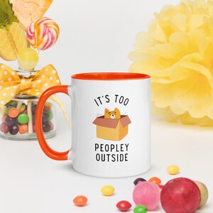 May include: White ceramic mug with an orange rim and handle. The mug has a cartoon cat in a box with the text "It's too peopley outside".