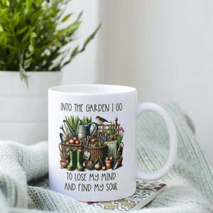 May include: White ceramic mug with a gardening themed design. The mug features a colorful illustration of gardening tools, plants, and a bird. The text on the mug reads "Into the garden I go to lose my mind and find my soul."