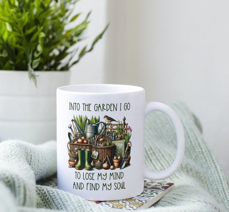 May include: White ceramic mug with a gardening themed design. The mug features a colorful illustration of gardening tools, plants, and a bird. The text on the mug reads "Into the garden I go to lose my mind and find my soul."