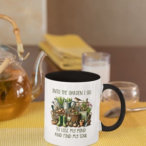 May include: White ceramic mug with a black rim and handle. The mug has a gardening themed design with the text "Into the garden I go to lose my mind and find my soul".