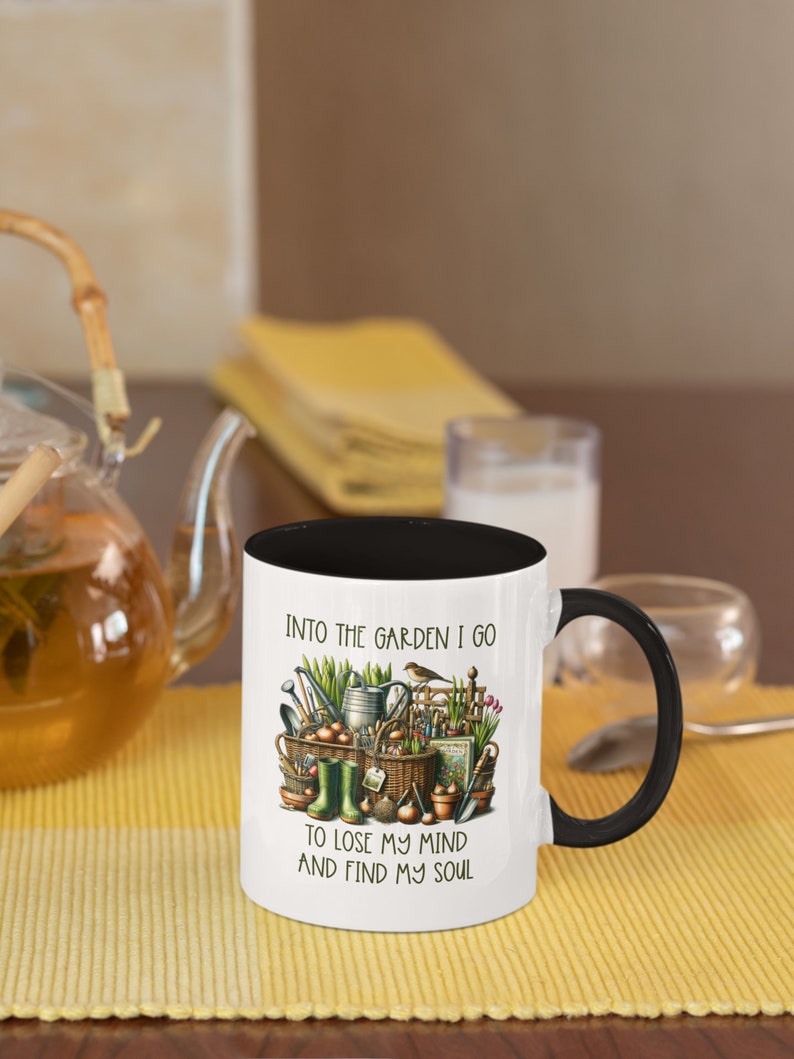 May include: White ceramic mug with a black rim and handle. The mug has a gardening themed design with the text "Into the garden I go to lose my mind and find my soul".