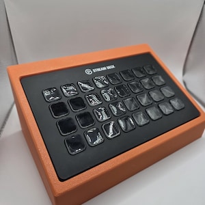 May include: A black Stream Deck control console with thirty-two square, translucent keys arranged in a grid. The device is housed within a textured, bright orange rectangular frame that sits at an angled position on a flat surface.