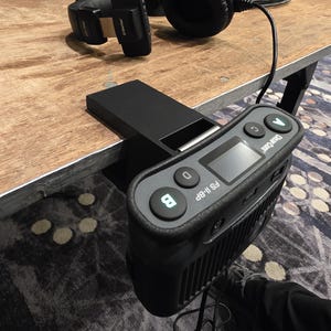 May include: A black Clear-Com FS II-BP beltpack intercom unit with illuminated buttons labeled A, B, C, and D, clamped to the edge of a table. A pair of black over-ear headphones is connected to the device by a black cable.