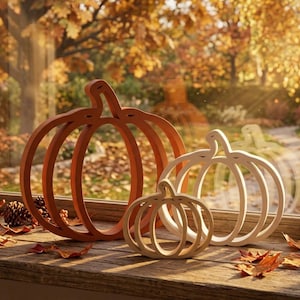 May include: Three open-frame pumpkin decorations of varying sizes arranged in a row. The largest is a deep orange, the medium is a light cream, and the smallest is a natural wood tone. Each pumpkin features a simple, hollowed-out silhouette design.