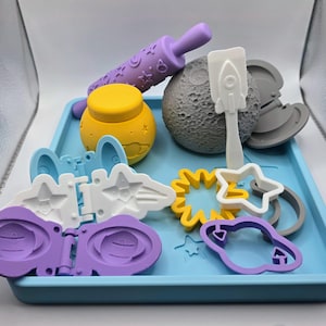 May include: A light blue rectangular tray containing various space-themed items including a purple rolling pin with star patterns, a yellow jar, a textured gray moon sphere, a white rocket spatula, and several plastic molds shaped like planets and stars.